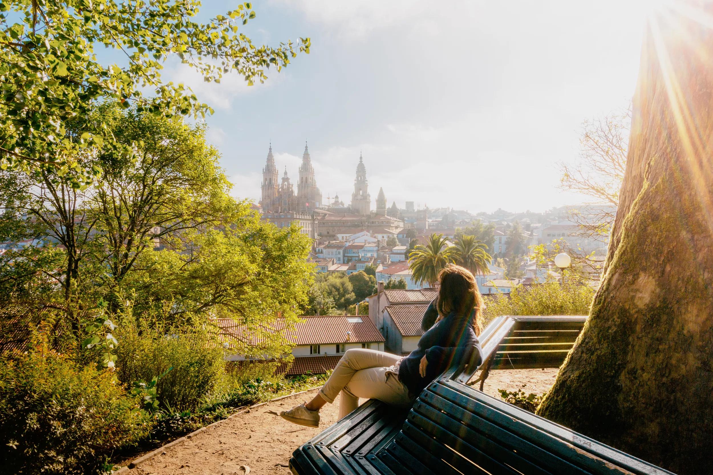 Woman on a park bench watching Santiago de Compostela Cathedral at sunrise, park and sunny sky. Galicia, Spain.