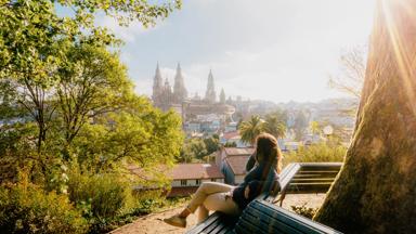 Woman on a park bench watching Santiago de Compostela Cathedral at sunrise, park and sunny sky. Galicia, Spain.
