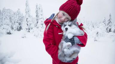 A girl dressed in red with a husky puppy standing in a winter wonderland.