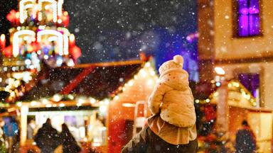 Little preschool girl sitting on shoulder of father on Christmas market in Germany. Happy toddler child and man observing traditional decorated pyramid. Happy family, bonding, love. Family xmas time
