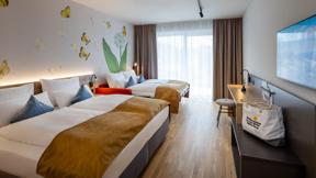 Sie sehen ein Family & Friends Zimmer für bis zu vier Personen mit Balkon, TV, komfortablem Boxspringbett, ausgezogener Schlafcouch, modernem Möbiliar und Badetasche am Zimmer.