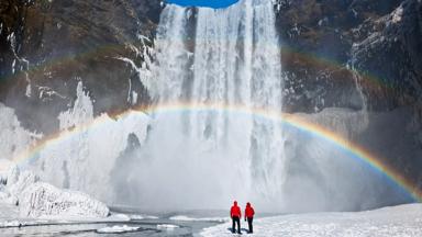 Couple under a rainbow by a large waterfall, winter. Skogafoss, Iceland.