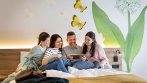Sie sehen eine lachende vierköpfige Familie gemütlich auf einem Bett sitzend beim gemeinsamen Entdecken von digitalen Inhalten auf einem Tablet im JUFA Hotel Bad Radkersburg