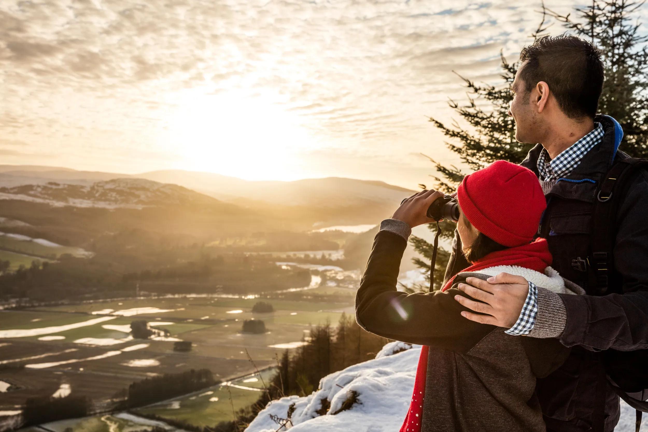 Two people, a man and young girl standing looking out over the scottish highland landscape in winter, one using binoculars.