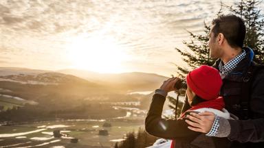 Two people, a man and young girl standing looking out over the scottish highland landscape in winter, one using binoculars.