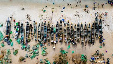 An overhead aerial shot of different colored boats on a sandy beach with the sea nearby