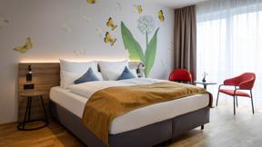 Sie sehen ein Doppelzimmer für zwei Personen im JUFA Hotel Bad Radkersburg mit Balkon, komfortablem Boxpringbett, modernem Möbiliar, gemütlicher Sitzecke mit Tisch und kreativer Wanddeko.