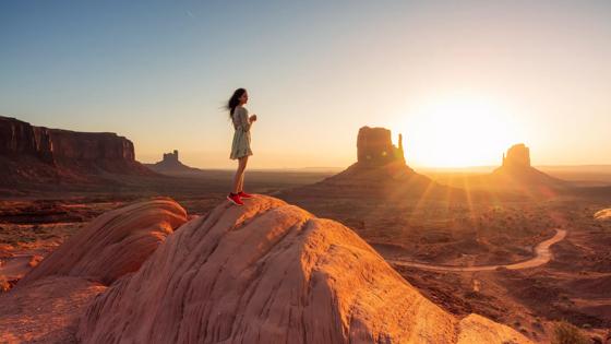 April 2022, a woman standing on the top of a rock with the sun rising beyond the Butte at Monument Valley, United States